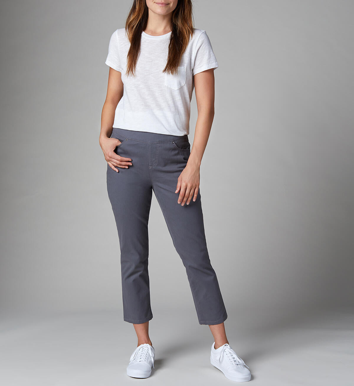 A person wearing gray Maya Crop-Jeans by JAG and a white shirt stands against a plain background. The high-rise jeans are cropped above the ankles, and the person is wearing white sneakers. One of their hands rests on their hip while the other hangs by their side.