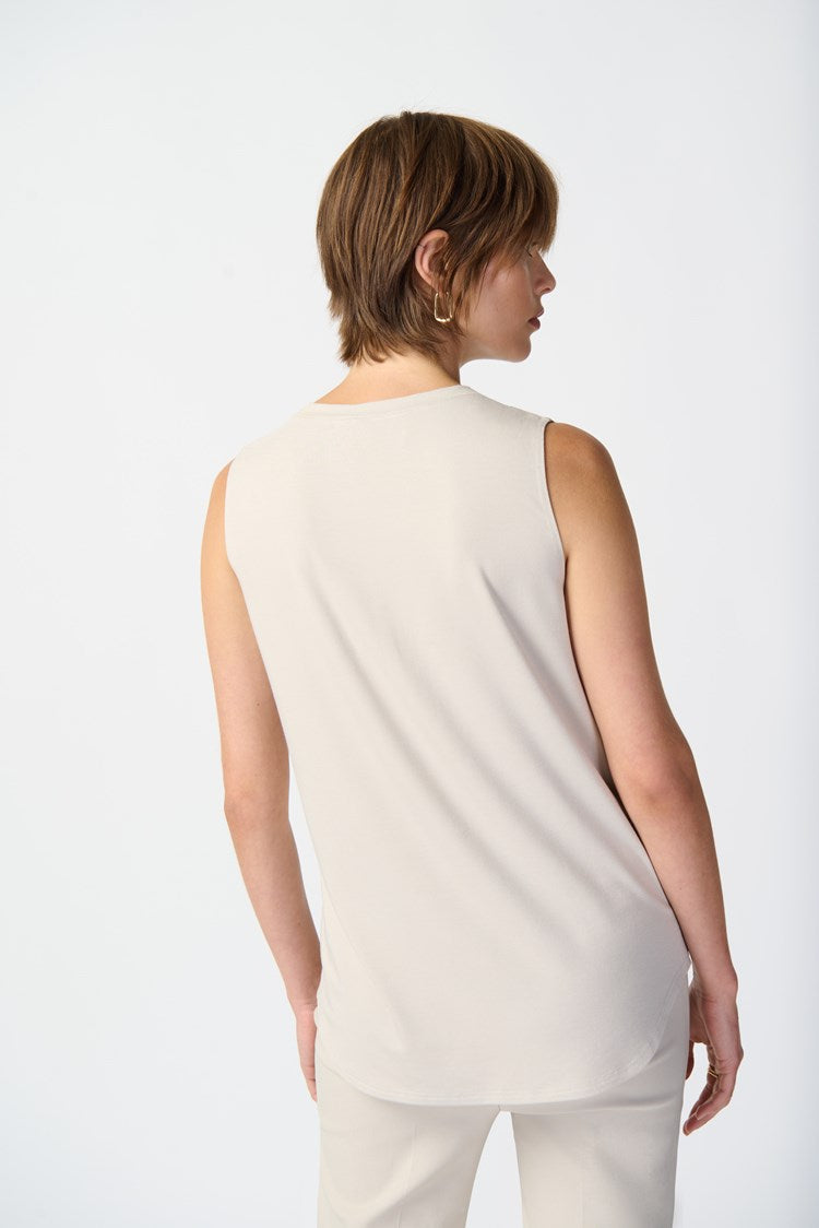 A person with short brown hair is standing against a plain white background. They are wearing a Bamboo Blend Sleeveless Jersey 241289 by Joseph Ribkoff, light gray slim-fit pants, and beige open-toe high-heeled shoes. Their right hand is in their pocket, and their left hand rests by their side.