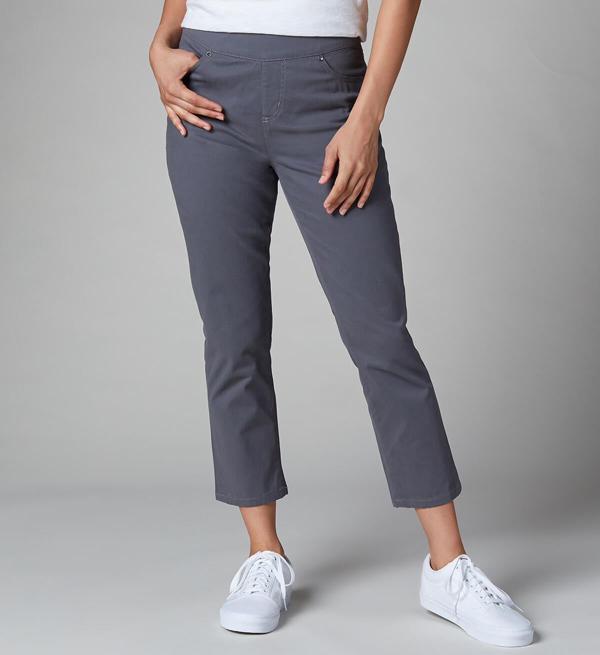 A person wearing gray Maya Crop-Jeans by JAG and a white shirt stands against a plain background. The high-rise jeans are cropped above the ankles, and the person is wearing white sneakers. One of their hands rests on their hip while the other hangs by their side.