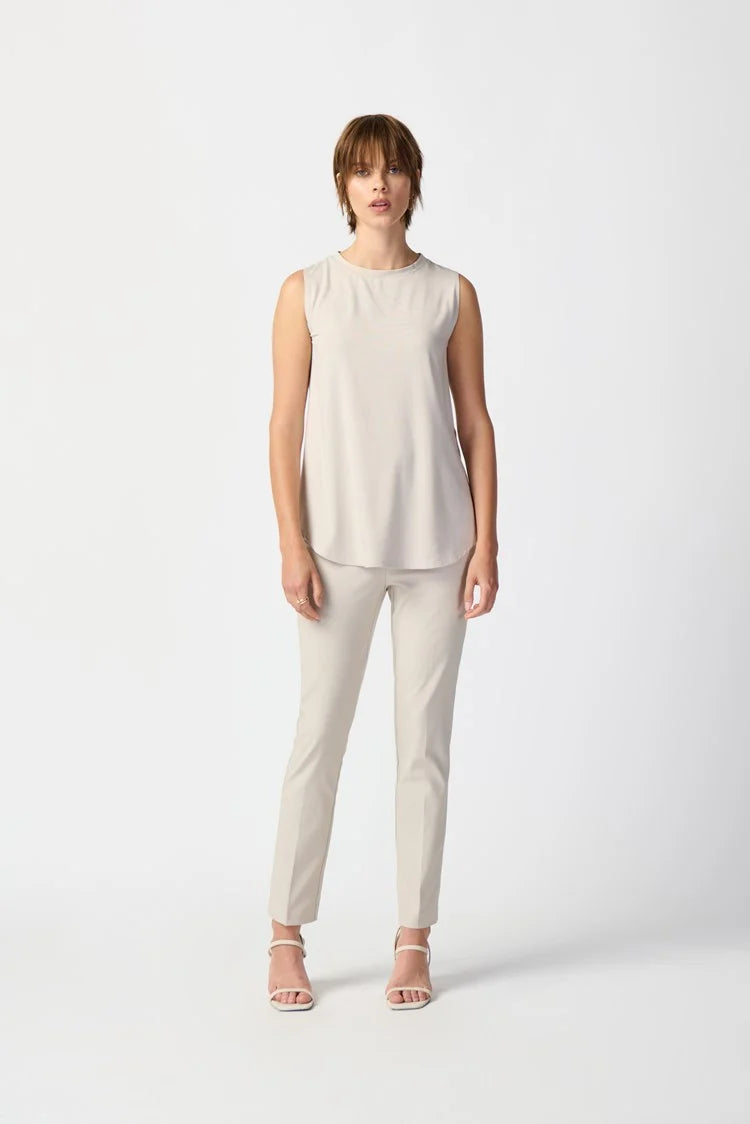 A person with short brown hair is standing against a plain white background. They are wearing a Bamboo Blend Sleeveless Jersey 241289 by Joseph Ribkoff, light gray slim-fit pants, and beige open-toe high-heeled shoes. Their right hand is in their pocket, and their left hand rests by their side.