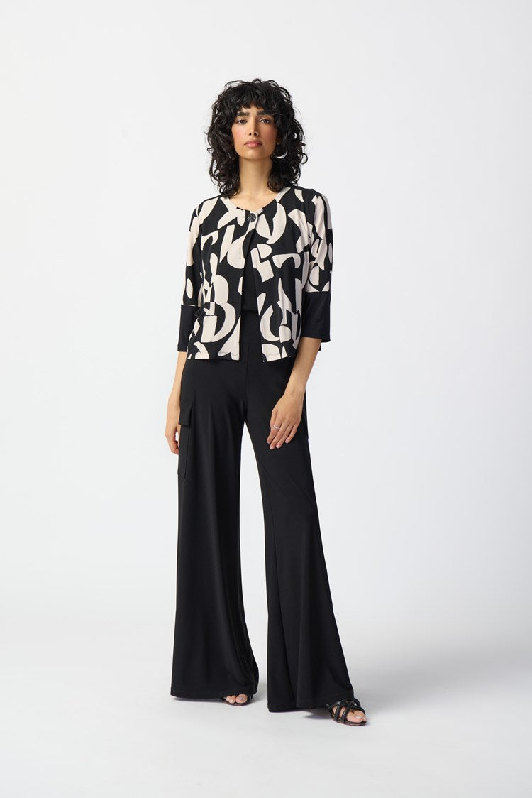 A woman with curly hair stands wearing the Joseph Ribkoff Abstract Print Silky Knit Jacket 241223, layered over a black top and wide-leg black pants. She poses confidently with one hand on her hip against a plain white background, complementing her outfit with black sandals.