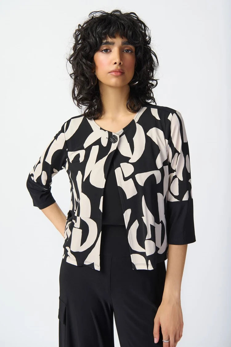 A woman with curly hair stands wearing the Joseph Ribkoff Abstract Print Silky Knit Jacket 241223, layered over a black top and wide-leg black pants. She poses confidently with one hand on her hip against a plain white background, complementing her outfit with black sandals.