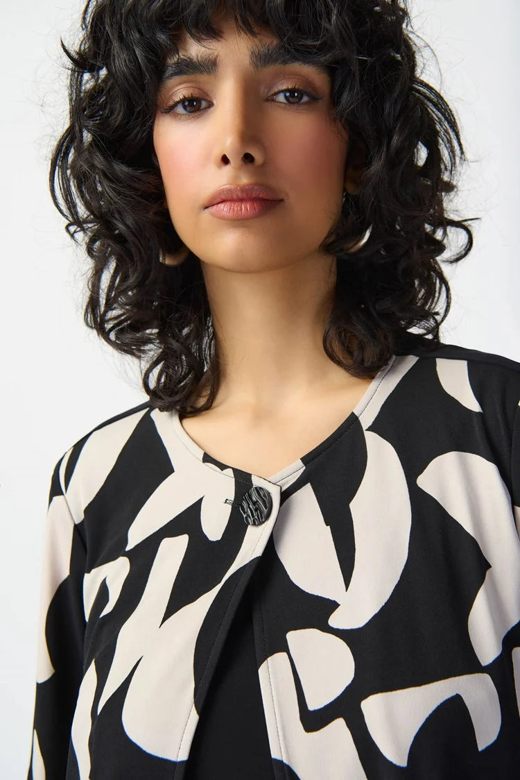 A woman with curly hair stands wearing the Joseph Ribkoff Abstract Print Silky Knit Jacket 241223, layered over a black top and wide-leg black pants. She poses confidently with one hand on her hip against a plain white background, complementing her outfit with black sandals.