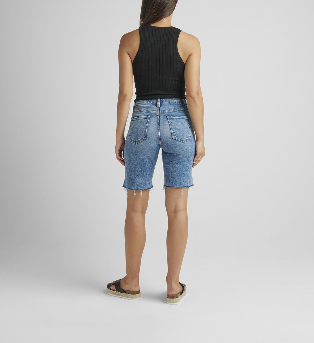 A person stands against a plain background, wearing a black sleeveless ribbed top and light blue City High Rise Shorts by JAG made from Power Stretch Denim with a frayed hem. They wear black platform sandals and have straight, long hair falling loose. Their right arm is relaxed by their side while the left arm is slightly bent.