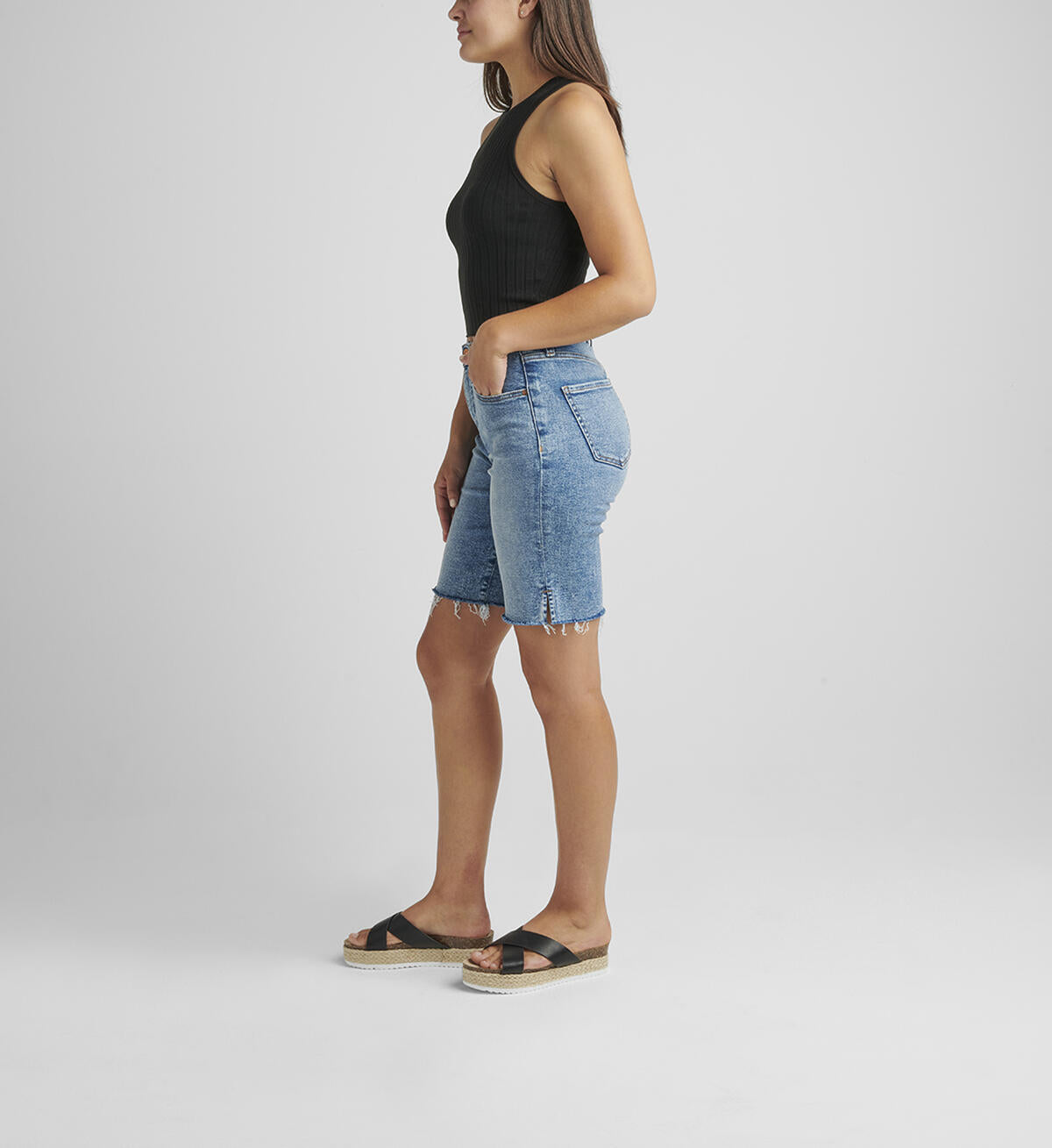 A person stands against a plain background, wearing a black sleeveless ribbed top and light blue City High Rise Shorts by JAG made from Power Stretch Denim with a frayed hem. They wear black platform sandals and have straight, long hair falling loose. Their right arm is relaxed by their side while the left arm is slightly bent.