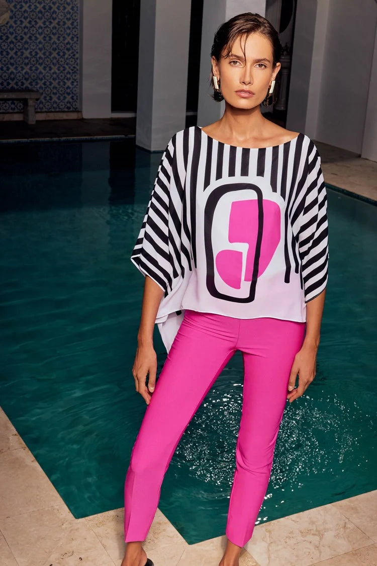 A woman stands confidently against a plain white background. She is wearing a stylish outfit consisting of the Abstract Print Georgette Poncho Top 241091 by Joseph Ribkoff, featuring black and white stripes with an abstract pink geometric design in the center, paired with white wide-leg pants. She completes the look with white heels.