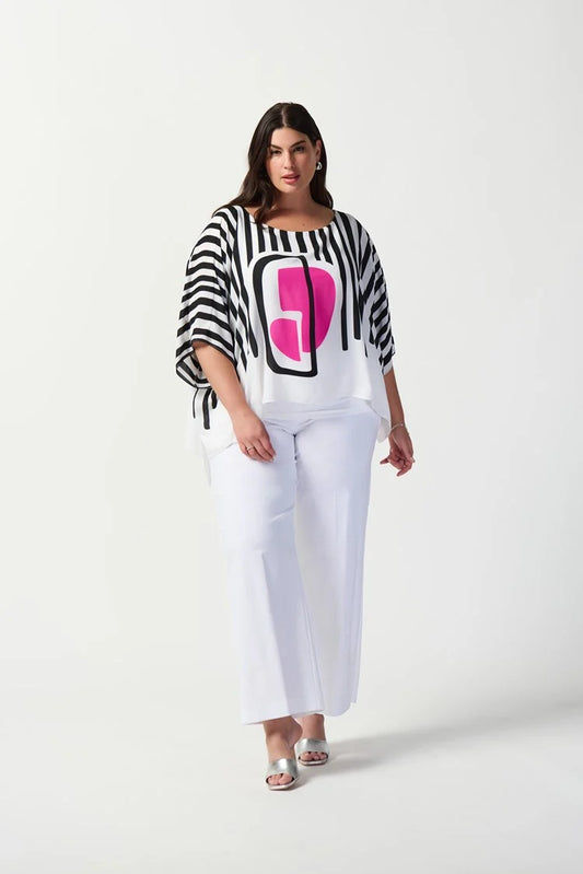 A woman stands confidently against a plain white background. She is wearing a stylish outfit consisting of the Abstract Print Georgette Poncho Top 241091 by Joseph Ribkoff, featuring black and white stripes with an abstract pink geometric design in the center, paired with white wide-leg pants. She completes the look with white heels.