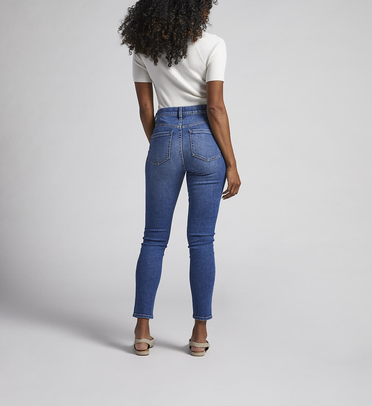 A person stands against a plain background, wearing a white short-sleeve ribbed turtleneck sweater, JAG Valentina Jeans that are high-waisted with an elastic waistband, and beige pointed-toe flats. They have curly hair cascading over their shoulders and their hands are relaxed by their sides.