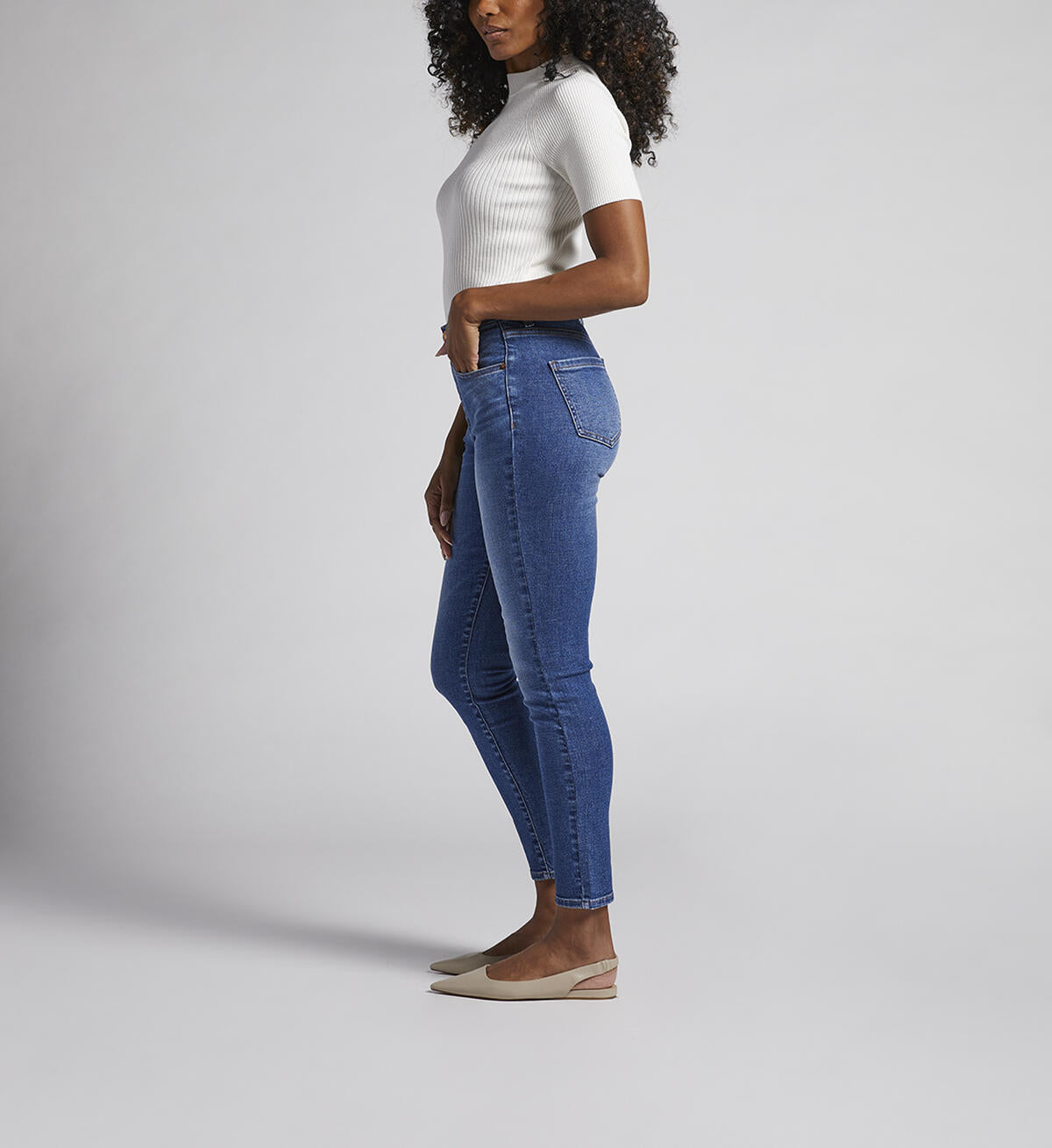 A person stands against a plain background, wearing a white short-sleeve ribbed turtleneck sweater, JAG Valentina Jeans that are high-waisted with an elastic waistband, and beige pointed-toe flats. They have curly hair cascading over their shoulders and their hands are relaxed by their sides.