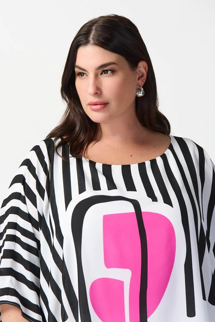 A woman stands confidently against a plain white background. She is wearing a stylish outfit consisting of the Abstract Print Georgette Poncho Top 241091 by Joseph Ribkoff, featuring black and white stripes with an abstract pink geometric design in the center, paired with white wide-leg pants. She completes the look with white heels.