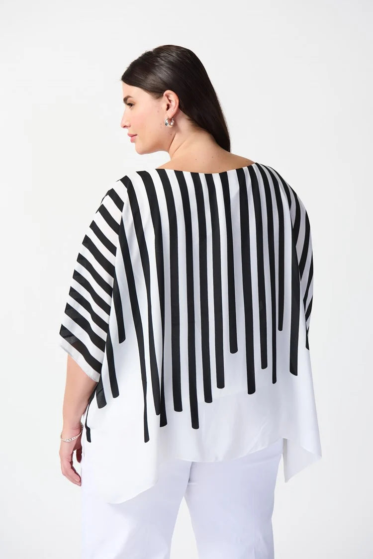 A woman stands confidently against a plain white background. She is wearing a stylish outfit consisting of the Abstract Print Georgette Poncho Top 241091 by Joseph Ribkoff, featuring black and white stripes with an abstract pink geometric design in the center, paired with white wide-leg pants. She completes the look with white heels.