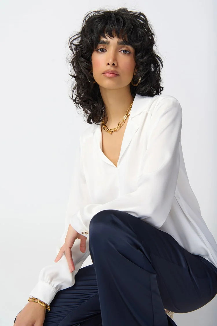 A person with curly black hair poses against a white background. They are wearing a loose-fitting Joseph Ribkoff Puff Sleeve Satin Top 241214 with a notch collar, dark pants, and gold jewelry including a necklace and bracelet. Their expression is neutral, and they are looking directly at the camera.