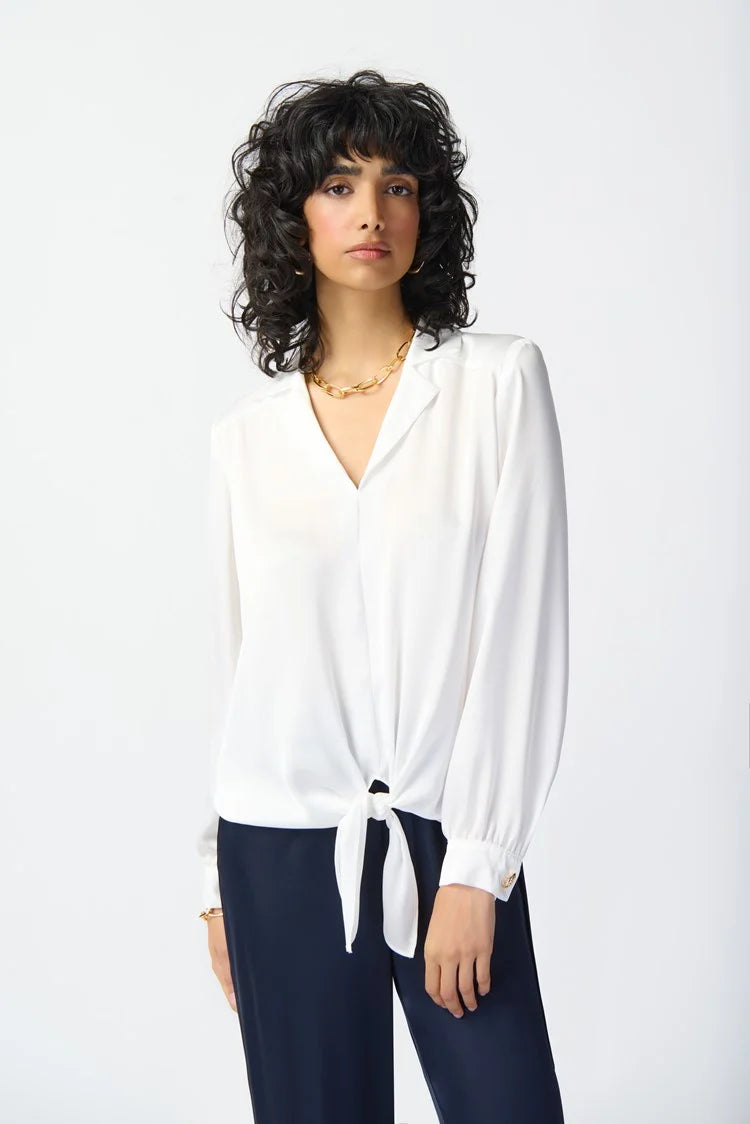 A person with curly black hair poses against a white background. They are wearing a loose-fitting Joseph Ribkoff Puff Sleeve Satin Top 241214 with a notch collar, dark pants, and gold jewelry including a necklace and bracelet. Their expression is neutral, and they are looking directly at the camera.