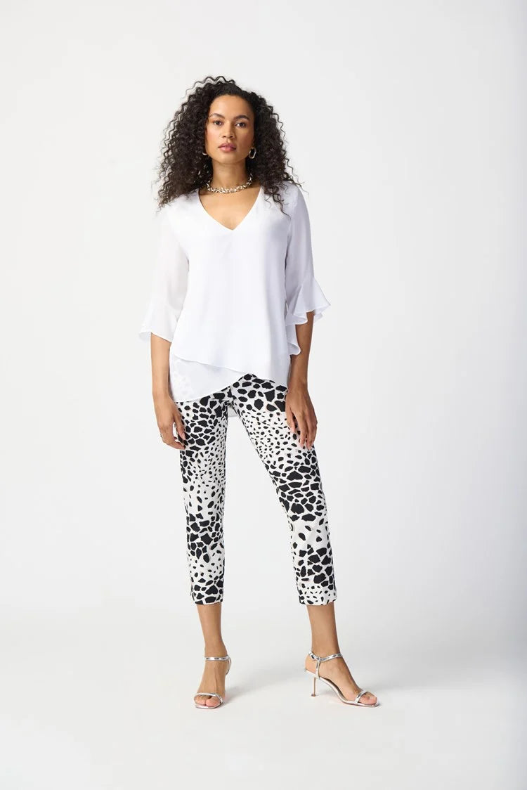 A woman with curly hair stands against a plain background, exuding feminine elegance in a Joseph Ribkoff Georgette Top With Ruffled Sleeves 241283, paired with three-quarter sleeves and cropped, patterned black and white pants. She completes the outfit with silver strappy heels and a simple necklace.