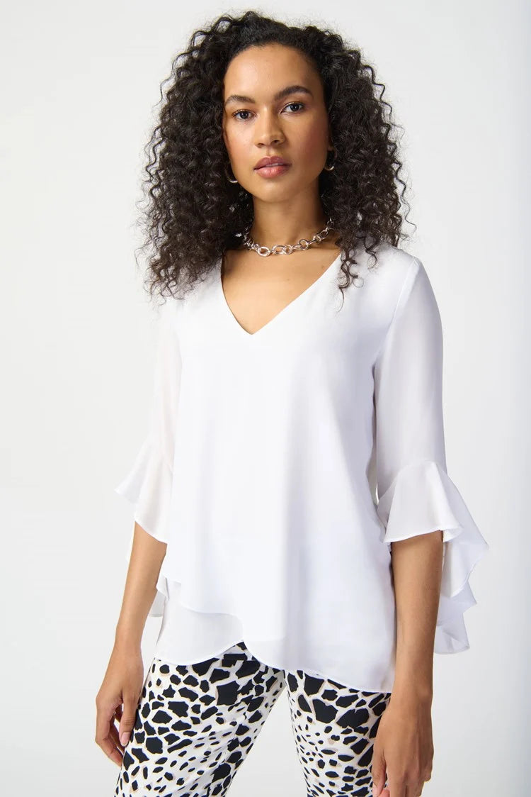 A woman with curly hair stands against a plain background, exuding feminine elegance in a Joseph Ribkoff Georgette Top With Ruffled Sleeves 241283, paired with three-quarter sleeves and cropped, patterned black and white pants. She completes the outfit with silver strappy heels and a simple necklace.