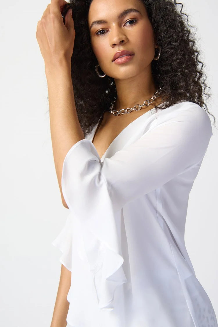 A woman with curly hair stands against a plain background, exuding feminine elegance in a Joseph Ribkoff Georgette Top With Ruffled Sleeves 241283, paired with three-quarter sleeves and cropped, patterned black and white pants. She completes the outfit with silver strappy heels and a simple necklace.