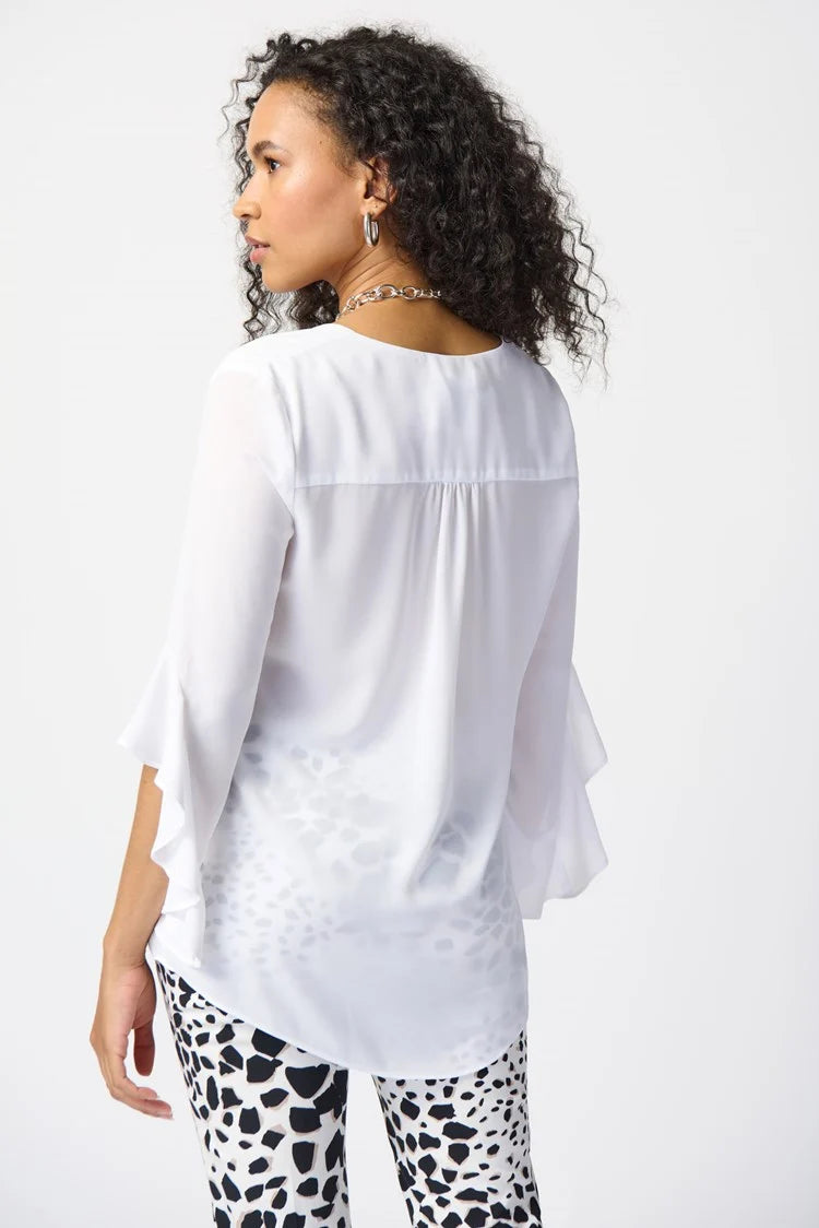 A woman with curly hair stands against a plain background, exuding feminine elegance in a Joseph Ribkoff Georgette Top With Ruffled Sleeves 241283, paired with three-quarter sleeves and cropped, patterned black and white pants. She completes the outfit with silver strappy heels and a simple necklace.