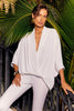 A person with short, slicked-back hair poses in front of lush green palm leaves. They are wearing the Joseph Ribkoff Woven Dolman Sleeve Wrap Top 241218, which features a deep V-neck and three-quarter dolman sleeves, paired with white slim-fit pants. They accessorize with large gold hoop earrings and a black and gold bracelet.