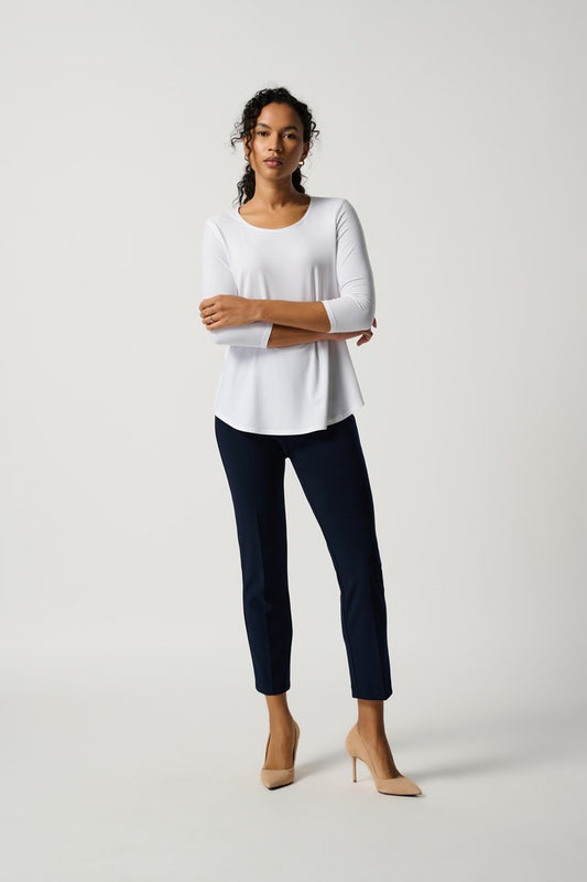 A person stands confidently in a simple, modern outfit consisting of a white, long-sleeved blouse, Joseph Ribkoff's Classic Cropped Pant in navy blue made from silky knit fabric, and beige high heels. They have a relaxed posture with one hand resting on their hip against the light gray background.