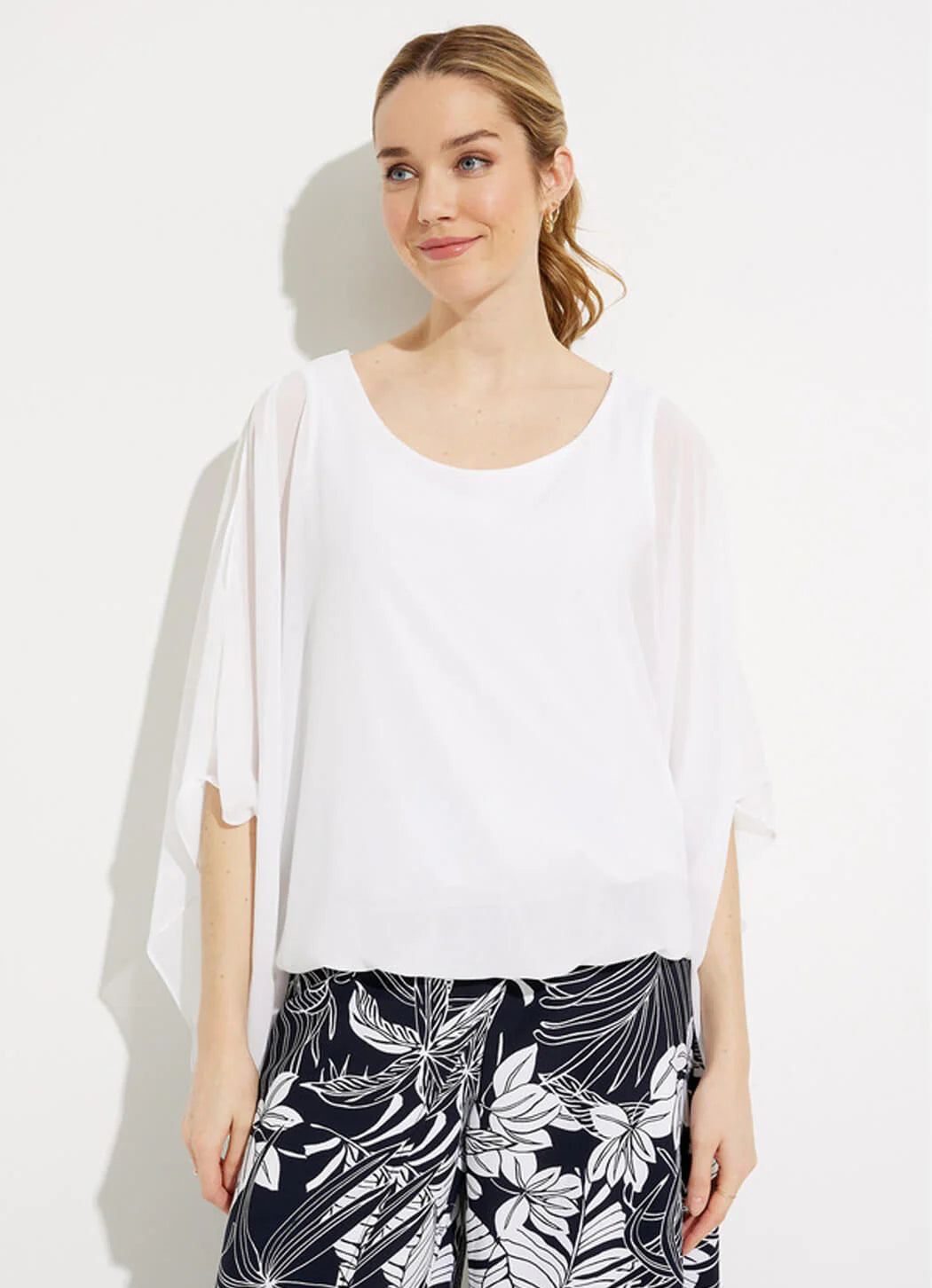 A person with light brown hair, styled in a ponytail, is standing against a plain white background. They are wearing the Joseph Ribkoff Two-Layer Chiffon Top 232131 by Joseph Ribkoff and black pants with a white floral pattern. They have a neutral expression and are looking slightly to the side.