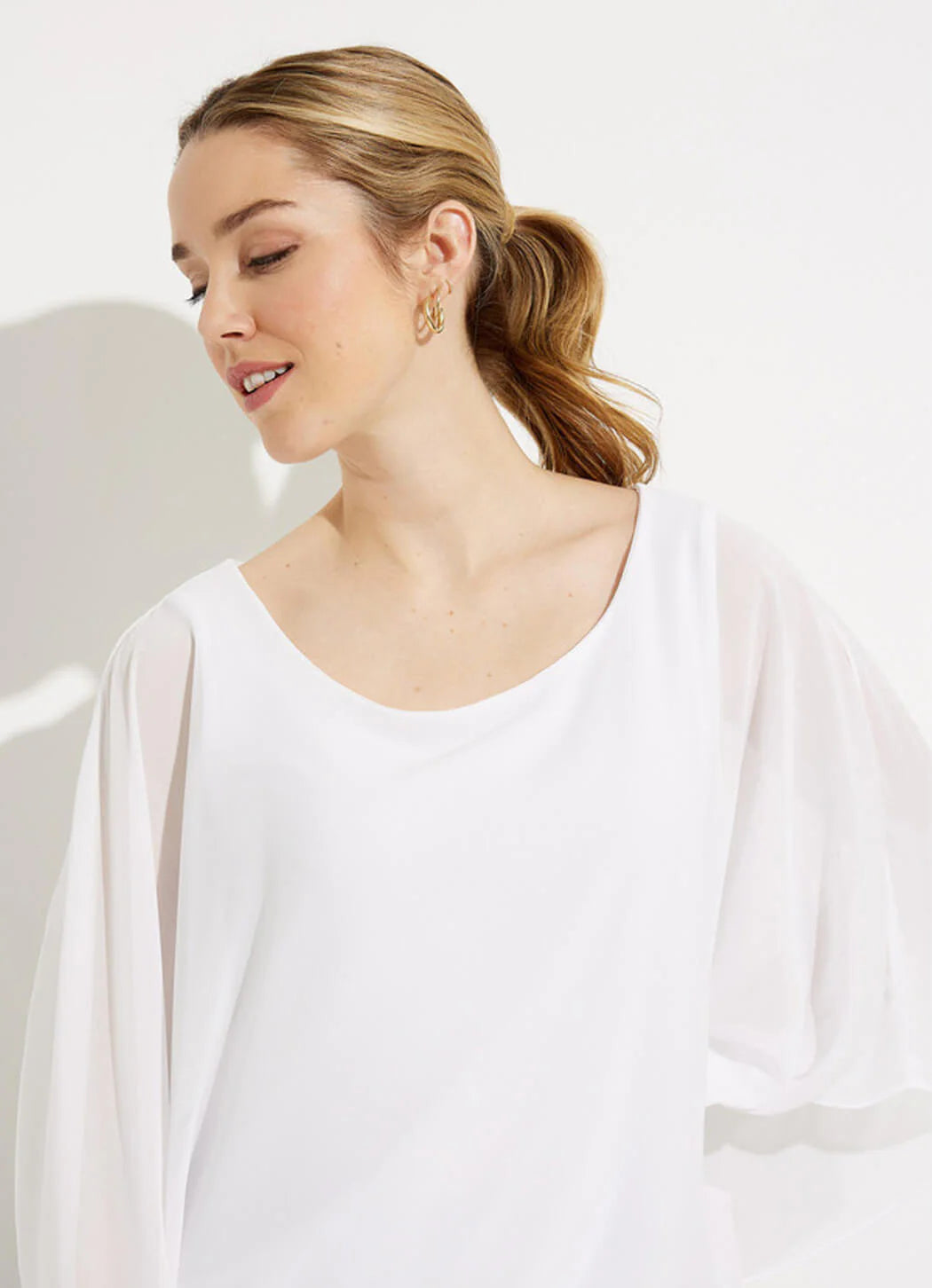 A person with light brown hair, styled in a ponytail, is standing against a plain white background. They are wearing the Joseph Ribkoff Two-Layer Chiffon Top 232131 by Joseph Ribkoff and black pants with a white floral pattern. They have a neutral expression and are looking slightly to the side.