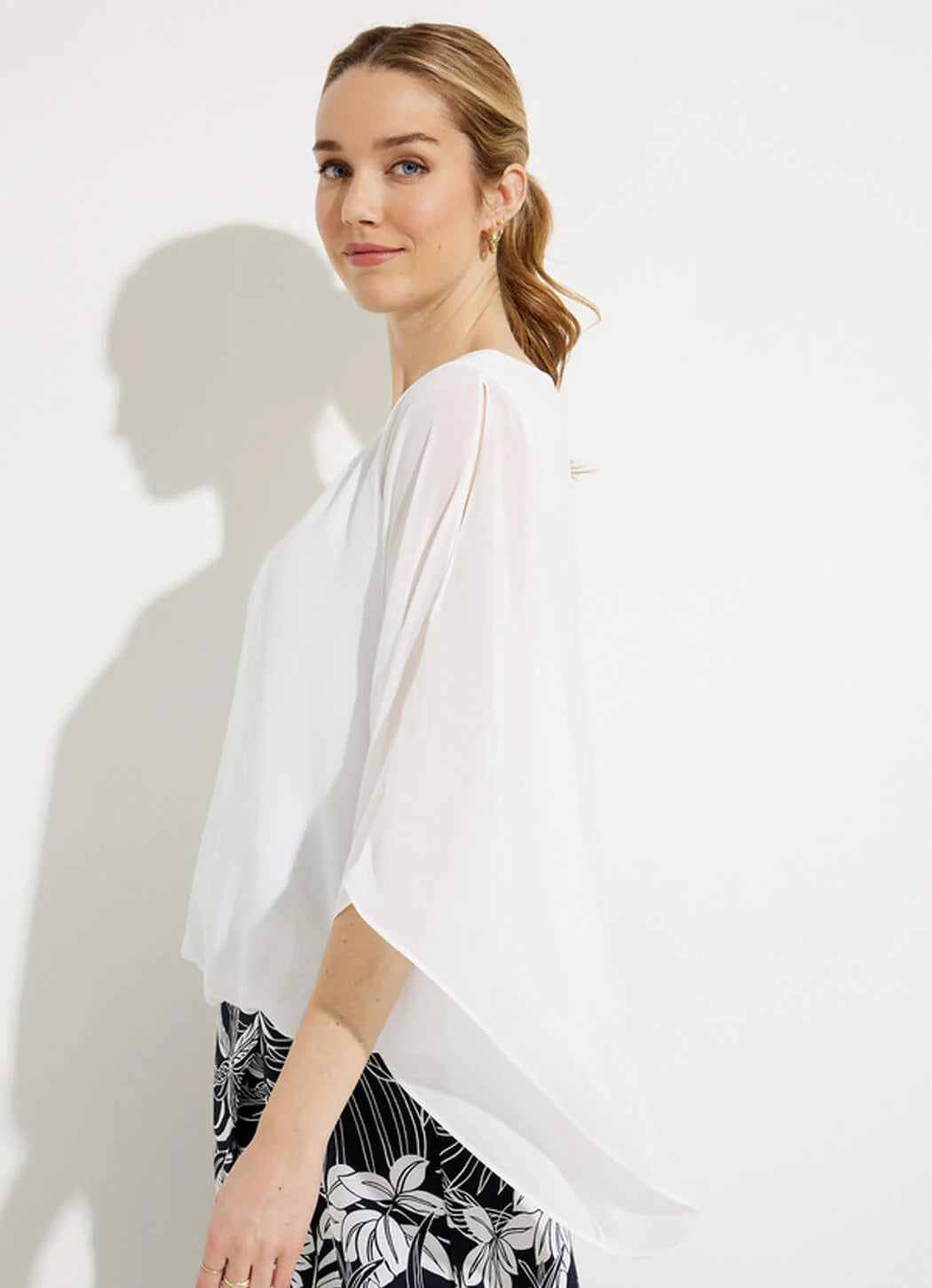 A person with light brown hair, styled in a ponytail, is standing against a plain white background. They are wearing the Joseph Ribkoff Two-Layer Chiffon Top 232131 by Joseph Ribkoff and black pants with a white floral pattern. They have a neutral expression and are looking slightly to the side.