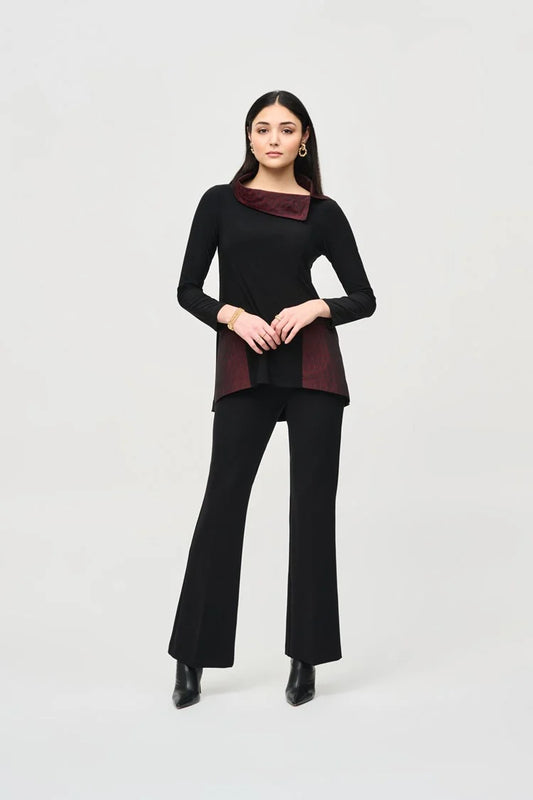 A woman with long dark hair stands confidently in a Joseph Ribkoff black outfit featuring a maroon collar and maroon panels at the waist. She wears the Silky Knit And Jacquard Memory Tunic paired with black pants and ankle boots. The background is plain light gray.