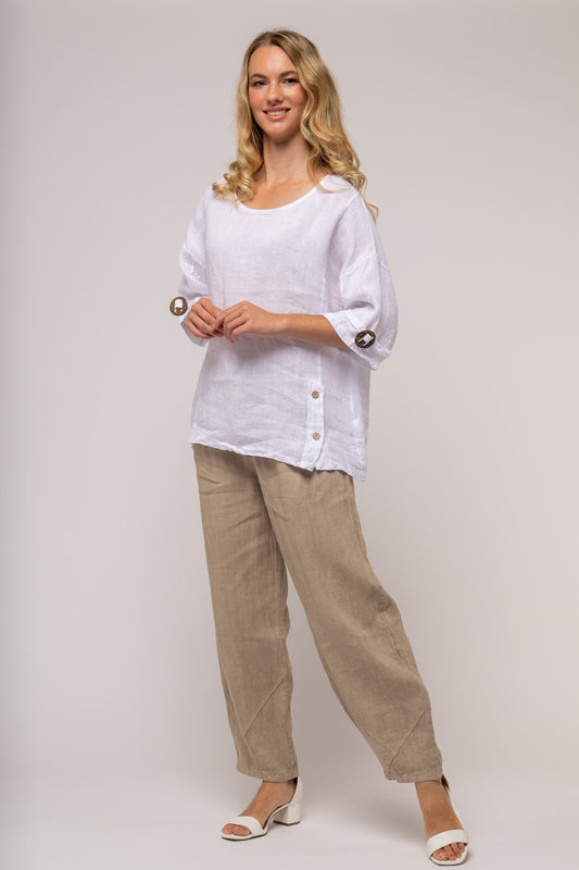 A smiling woman with wavy blonde hair is standing and modeling a casual outfit from LinenLuv's collection. She is wearing the LINEN LUV - French Linen Top TP1327-11 in Bianca, which features button details on the sleeves and sides and has a loose-fitting design. She pairs this white top with beige, wide-leg pants and complements the ensemble with white open-toe sandals. The background is plain.