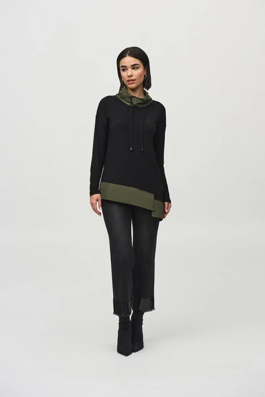 A woman stands against a plain background wearing the Joseph Ribkoff Silky Knit And Memory Asymmetrical Tunic, a black long-sleeve top with a green hood and an asymmetrical hem detail. The boxy tunic is paired with black frayed-hem jeans and black ankle boots. She has dark hair and a neutral expression.