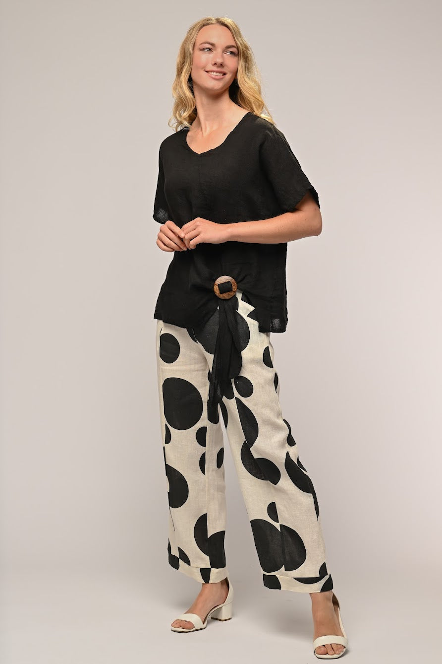 A smiling woman with long blonde hair, wearing a LinenLuv LINEN LUV - French Linen Top TP1157-74 in Nero and white wide-legged pants with black abstract circular patterns. She has on white open-toed sandals and a black belt with a large circular buckle. The background is plain light gray.