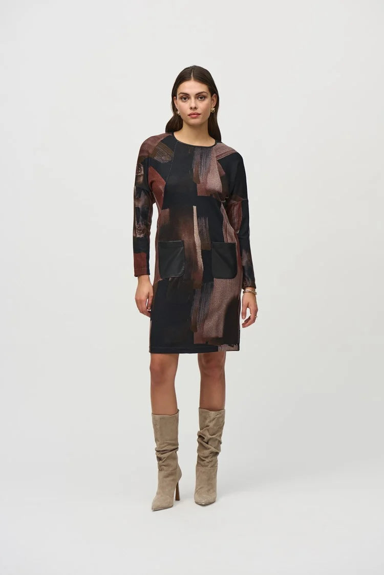 A woman with long dark hair stands against a plain background, wearing the Joseph Ribkoff Sweater Knit Abstract Print Dress 244123. This long-sleeve dress features an abstract print in black, brown, and beige tones. She pairs it with knee-high suede beige boots and has a neutral facial expression and relaxed posture.