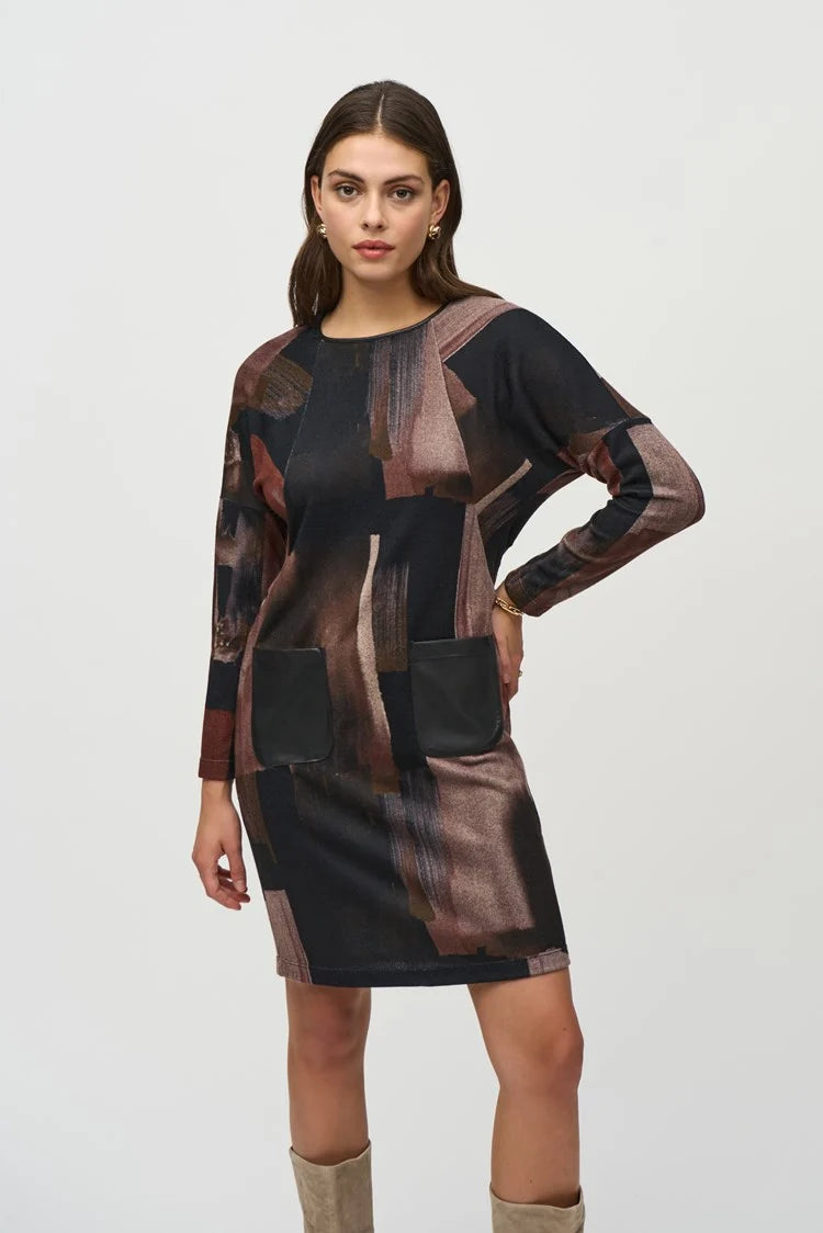 A woman with long dark hair stands against a plain background, wearing the Joseph Ribkoff Sweater Knit Abstract Print Dress 244123. This long-sleeve dress features an abstract print in black, brown, and beige tones. She pairs it with knee-high suede beige boots and has a neutral facial expression and relaxed posture.