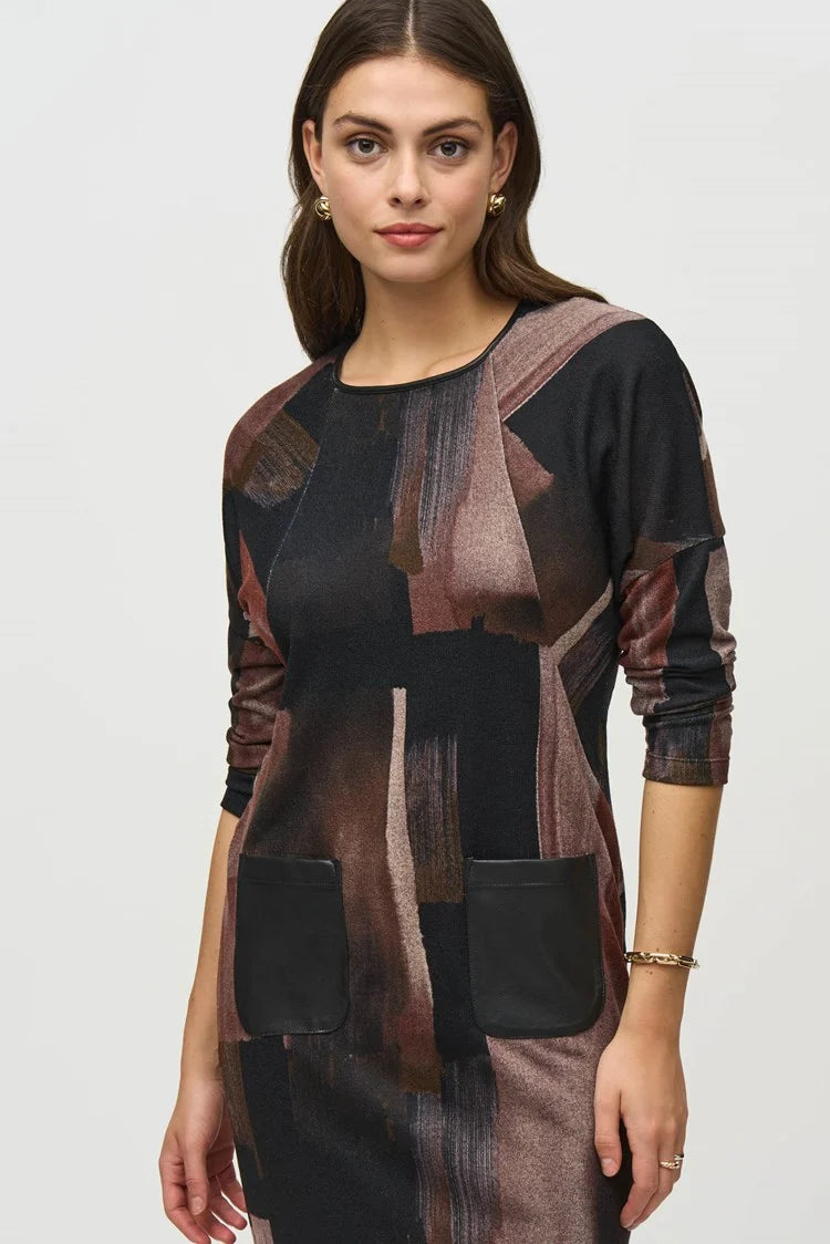 A woman with long dark hair stands against a plain background, wearing the Joseph Ribkoff Sweater Knit Abstract Print Dress 244123. This long-sleeve dress features an abstract print in black, brown, and beige tones. She pairs it with knee-high suede beige boots and has a neutral facial expression and relaxed posture.