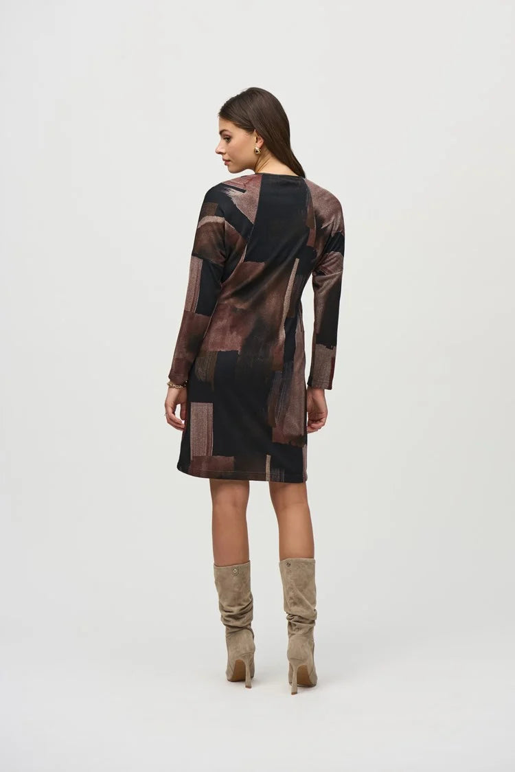 A woman with long dark hair stands against a plain background, wearing the Joseph Ribkoff Sweater Knit Abstract Print Dress 244123. This long-sleeve dress features an abstract print in black, brown, and beige tones. She pairs it with knee-high suede beige boots and has a neutral facial expression and relaxed posture.