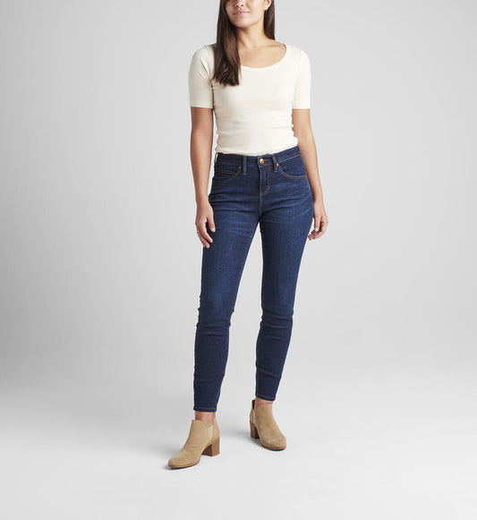 A woman with shoulder-length hair stands against a plain background. She is wearing a cream-colored short-sleeve top, Cecilia Mid Rise Skinny Jeans by JAG in Night Breeze, and light brown heeled ankle boots. Her right hand is relaxed by her side while her left hand is slightly raised.