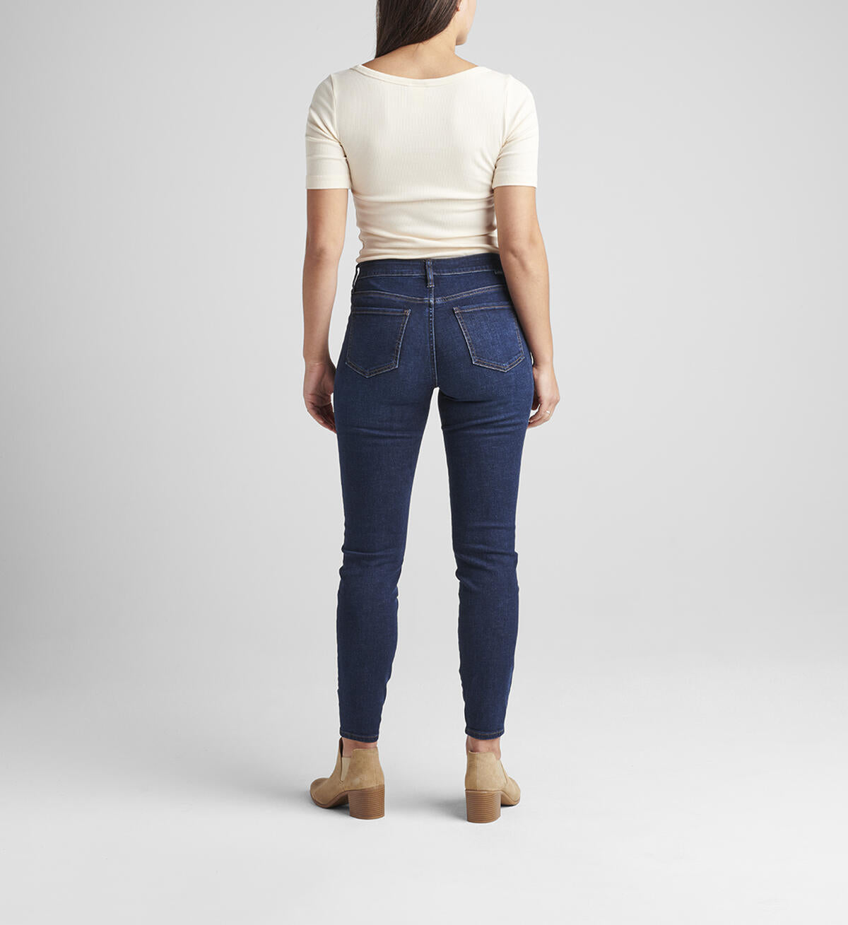 A woman with shoulder-length hair stands against a plain background. She is wearing a cream-colored short-sleeve top, Cecilia Mid Rise Skinny Jeans by JAG in Night Breeze, and light brown heeled ankle boots. Her right hand is relaxed by her side while her left hand is slightly raised.