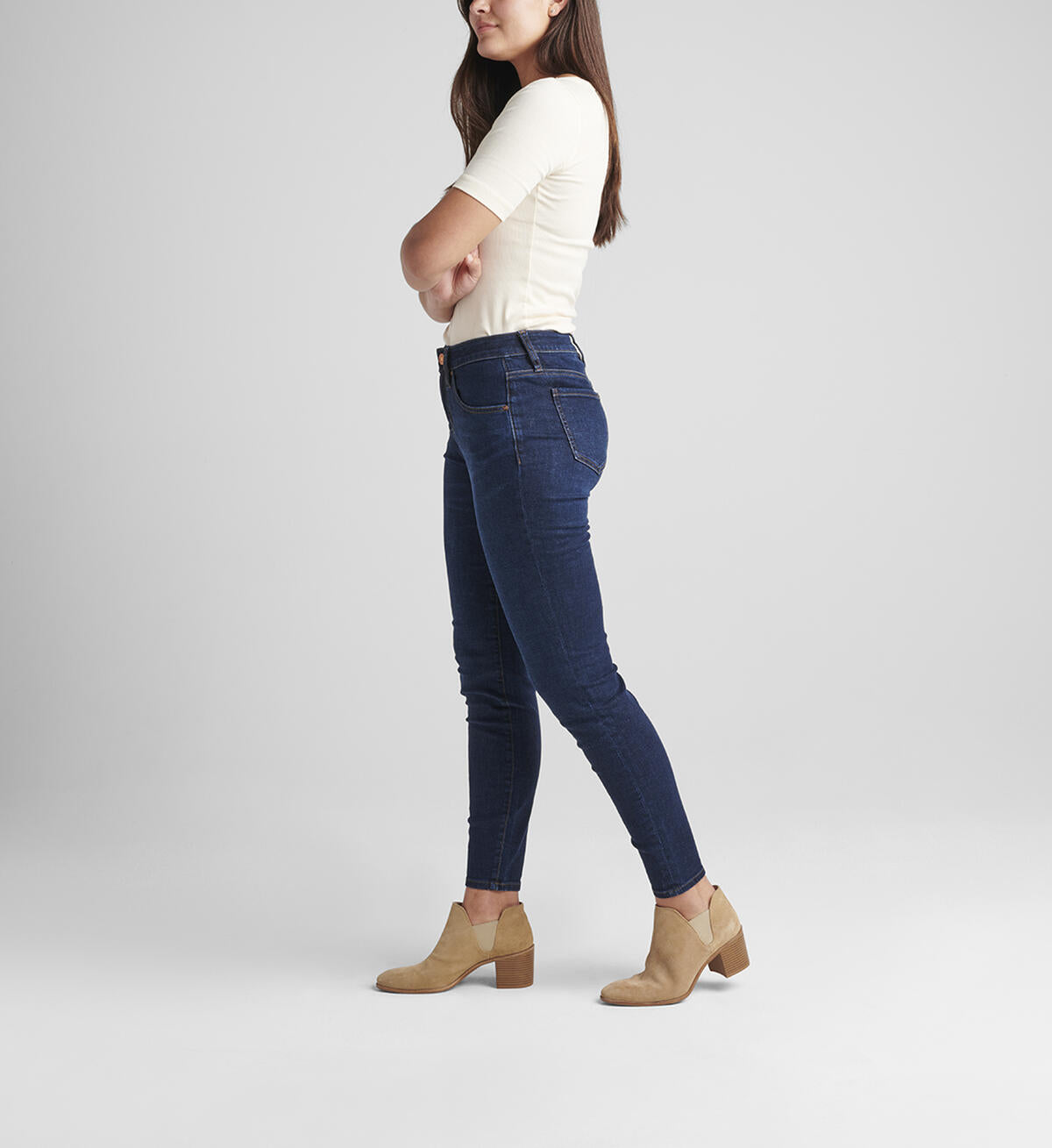 A woman with shoulder-length hair stands against a plain background. She is wearing a cream-colored short-sleeve top, Cecilia Mid Rise Skinny Jeans by JAG in Night Breeze, and light brown heeled ankle boots. Her right hand is relaxed by her side while her left hand is slightly raised.