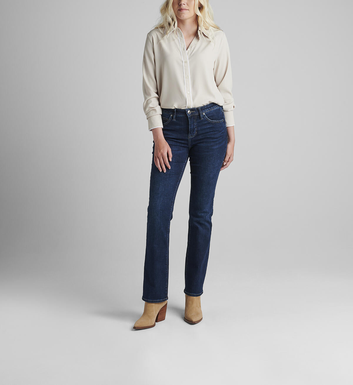 A person with shoulder-length blonde hair wearing a white long-sleeve blouse and JAG's dark blue Eloise Bootcut jeans in Night Breeze stands against a plain, light gray background. They have beige heeled ankle boots on and their hands are relaxed by their sides, effortlessly moving with ease thanks to the comfort stretch denim.