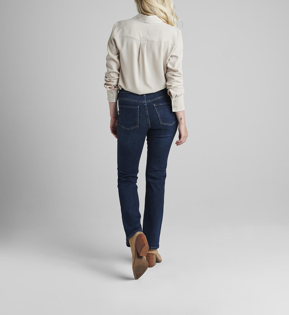 A person with shoulder-length blonde hair wearing a white long-sleeve blouse and JAG's dark blue Eloise Bootcut jeans in Night Breeze stands against a plain, light gray background. They have beige heeled ankle boots on and their hands are relaxed by their sides, effortlessly moving with ease thanks to the comfort stretch denim.