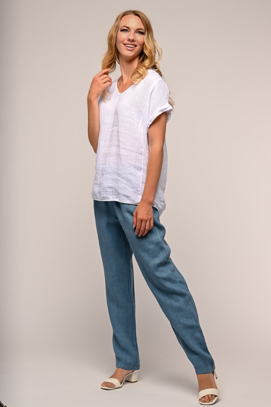 A blonde woman poses against a neutral background, smiling while looking slightly to the side. She is wearing a 100% linen white short-sleeve LINEN LUV - French Linen Top TP864-11, blue relaxed-fit pants, and white open-toe heels. One hand is touching her hair, the other rests by her side.