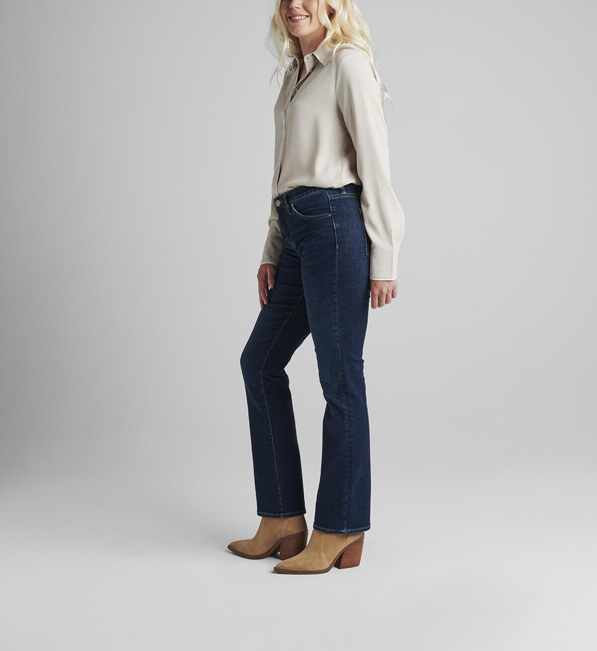 A person with shoulder-length blonde hair wearing a white long-sleeve blouse and JAG's dark blue Eloise Bootcut jeans in Night Breeze stands against a plain, light gray background. They have beige heeled ankle boots on and their hands are relaxed by their sides, effortlessly moving with ease thanks to the comfort stretch denim.