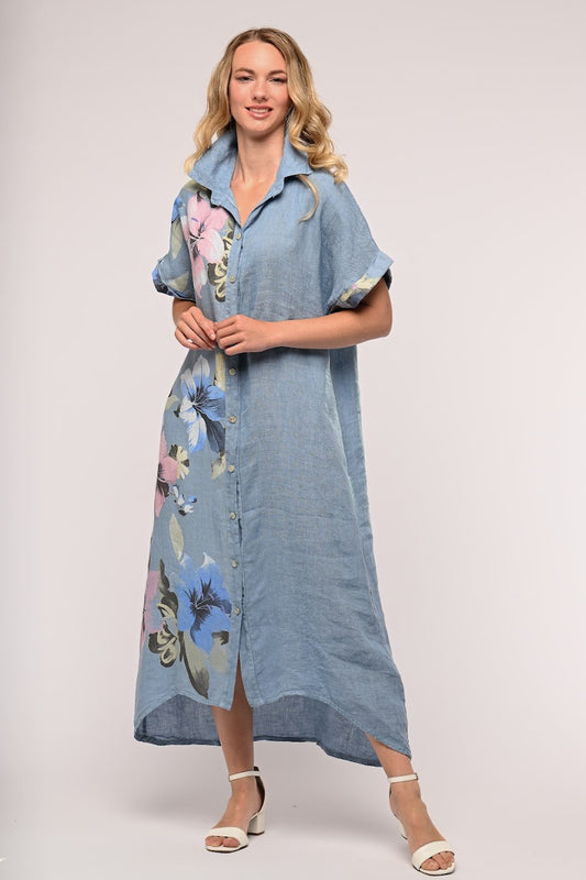 A woman with long blonde hair is wearing a blue, button-up, short-sleeve maxi dress with colorful floral patterns on one side. This "LINEN LUV - French Linen Dress TP1166-56 - Jeans" from LinenLuv pairs perfectly with her white sandals. She poses against a plain light grey background, resting her hands together at her waist and smiling.