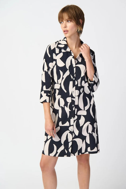 A woman stands against a plain white background, wearing the Joseph Ribkoff Abstract Print Stretch Poplin Trapeze Dress 241000. The dress features a long-sleeved, black and white abstract print design with a collar and buttons down the front. She has short, light brown hair and is accessorized with a pair of gold earrings.