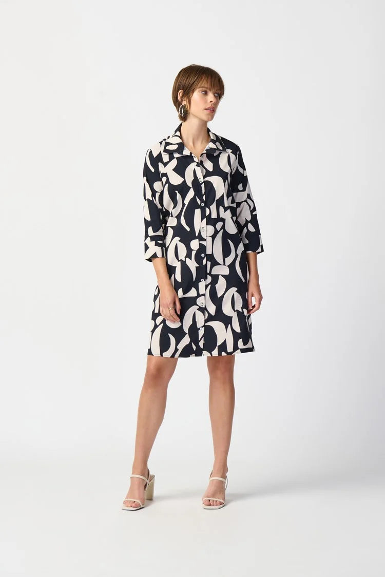 A woman stands against a plain white background, wearing the Joseph Ribkoff Abstract Print Stretch Poplin Trapeze Dress 241000. The dress features a long-sleeved, black and white abstract print design with a collar and buttons down the front. She has short, light brown hair and is accessorized with a pair of gold earrings.