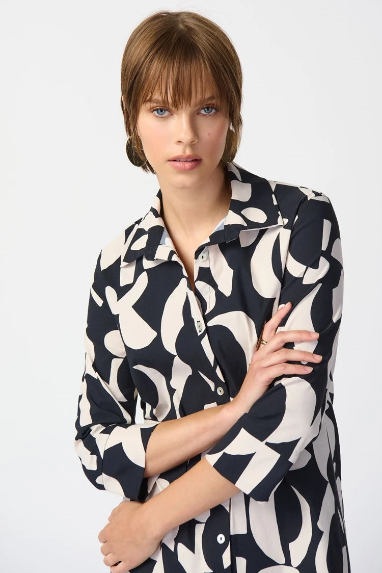 A woman stands against a plain white background, wearing the Joseph Ribkoff Abstract Print Stretch Poplin Trapeze Dress 241000. The dress features a long-sleeved, black and white abstract print design with a collar and buttons down the front. She has short, light brown hair and is accessorized with a pair of gold earrings.