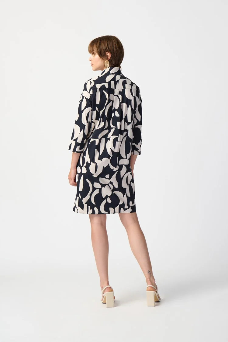 A woman stands against a plain white background, wearing the Joseph Ribkoff Abstract Print Stretch Poplin Trapeze Dress 241000. The dress features a long-sleeved, black and white abstract print design with a collar and buttons down the front. She has short, light brown hair and is accessorized with a pair of gold earrings.