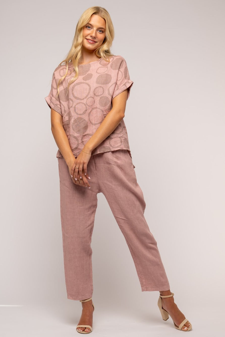 A person with long blonde hair is wearing a pink, short-sleeved LINEN O - French Linen Top TP1238-90 by LinenO, featuring a circular pattern, along with matching pink pants. They are standing and smiling, with their hands loosely clasped in front of them against a plain gray background.