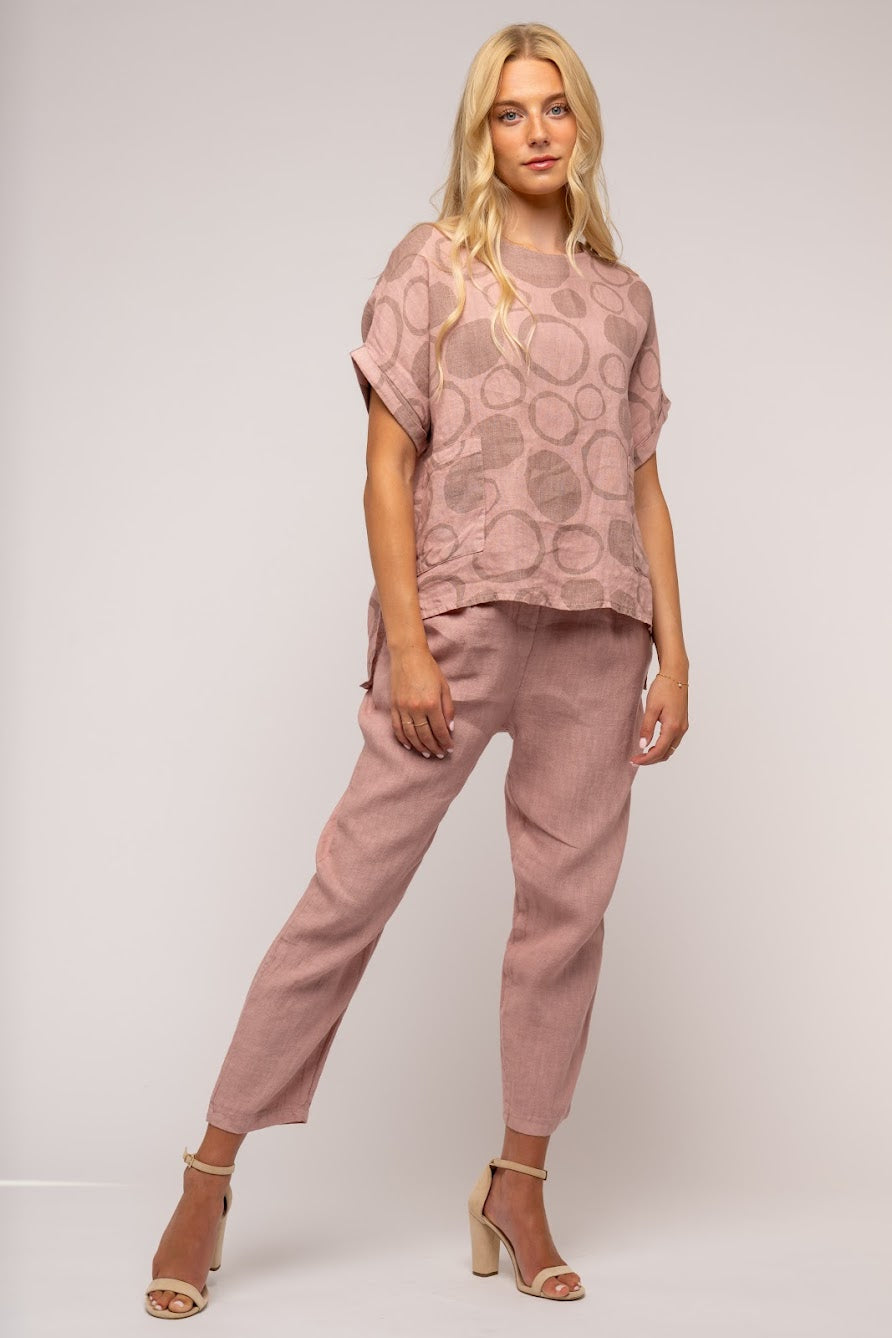 A person with long blonde hair is wearing a pink, short-sleeved LINEN O - French Linen Top TP1238-90 by LinenO, featuring a circular pattern, along with matching pink pants. They are standing and smiling, with their hands loosely clasped in front of them against a plain gray background.