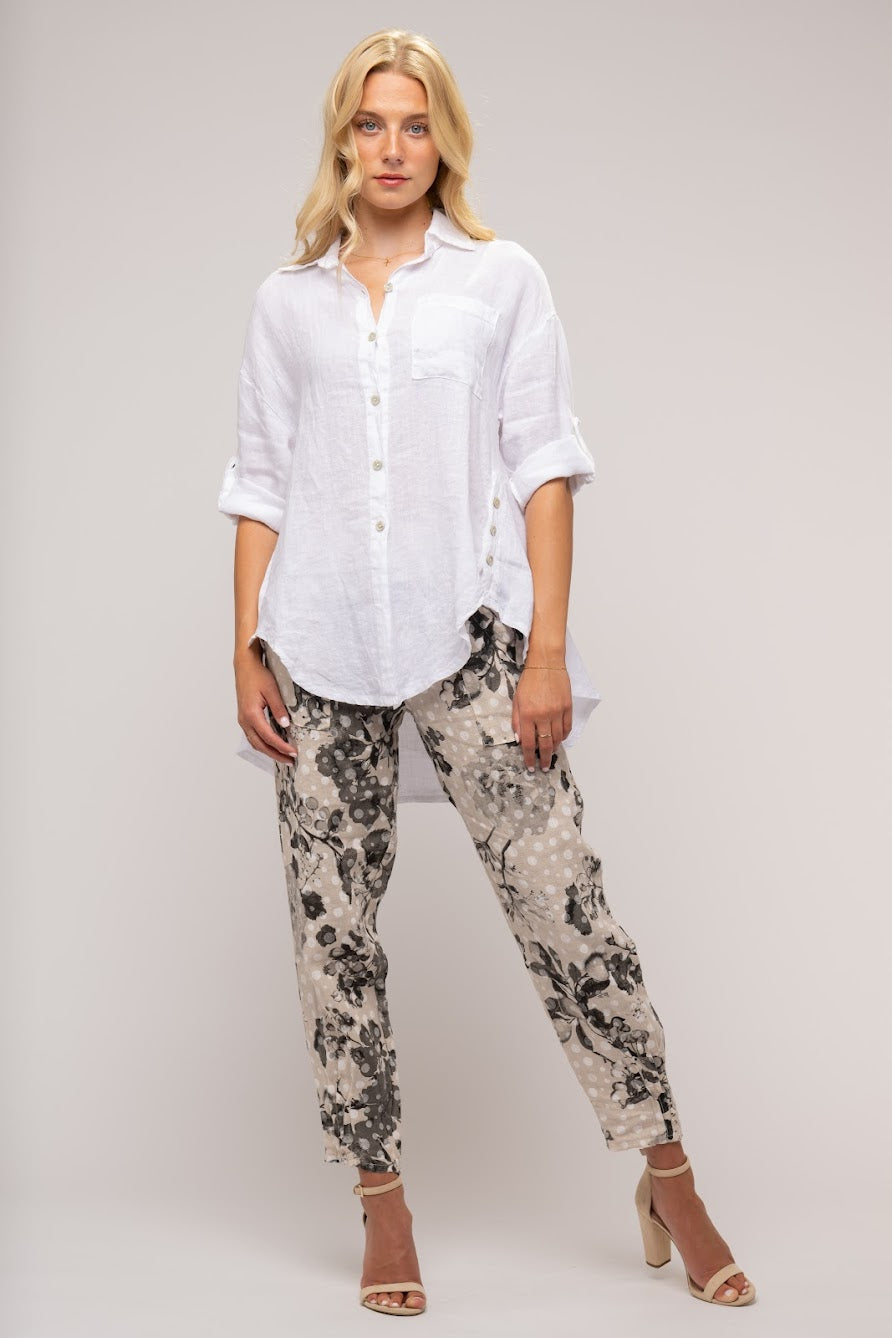 A blonde woman stands against a plain background, wearing the LINEN O - French Linen Shirt TP1154-11 by LinenO, a white button-up shirt with three-quarter sleeves crafted in Europe, and printed gray and black pants. She is also wearing beige high-heeled sandals. She faces slightly to the side, with a neutral expression.