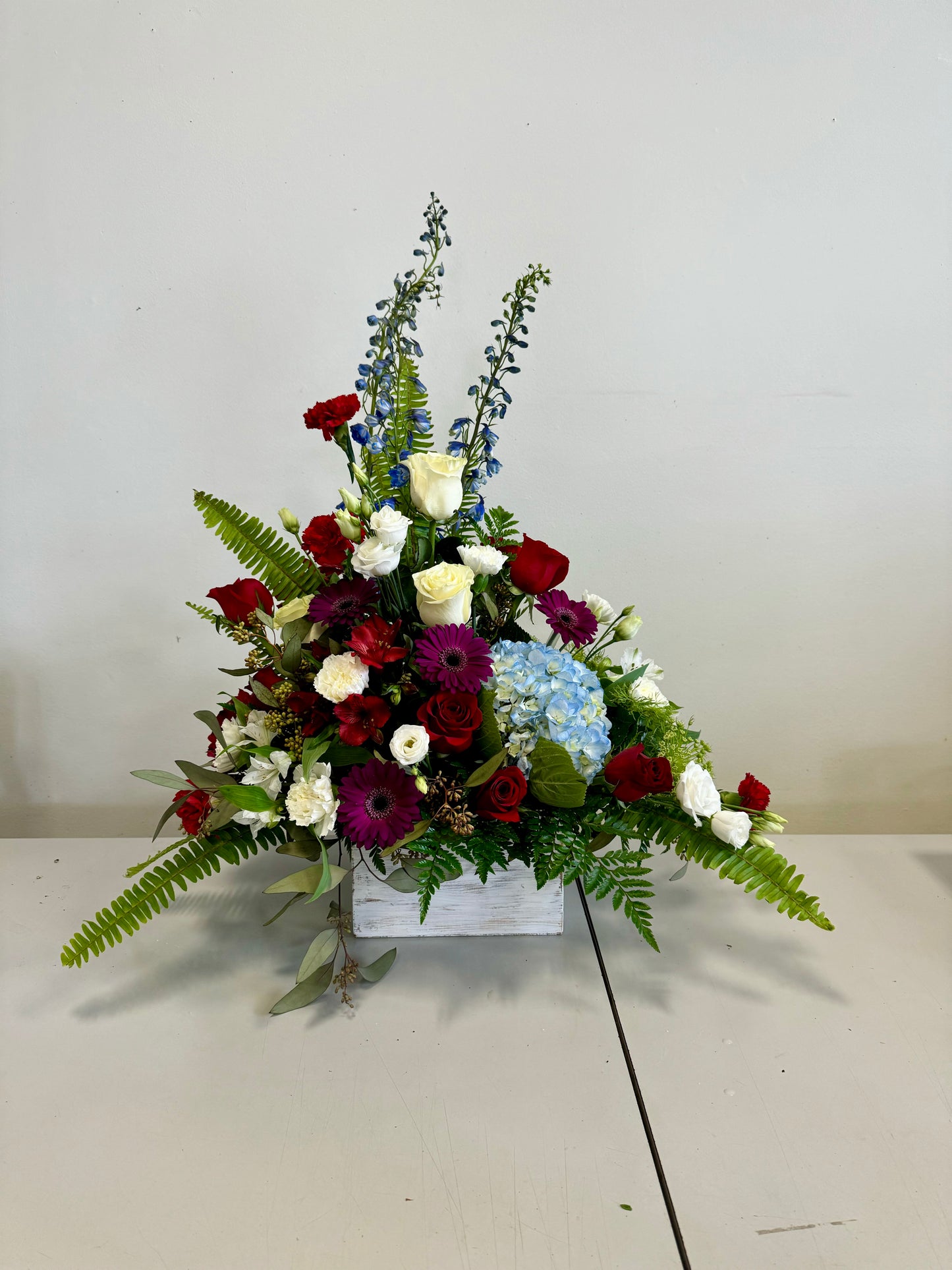 The Home Tribute by Posies Flowers & Fashion is a vibrant floral arrangement featuring red roses, white hydrangeas, purple blooms, and blue hydrangeas, complemented by gerbera daisies and green ferns. The flowers are elegantly arranged in a white wooden box and displayed on a gray table with a plain background.